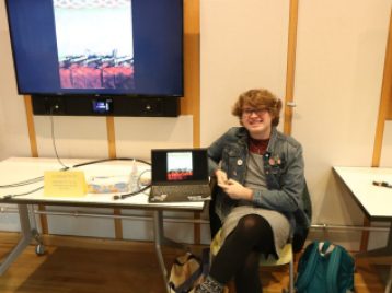 August Mashburn sits beside a laptop and large monitor displaying their project, "Glitched Emotions during Quarantine."